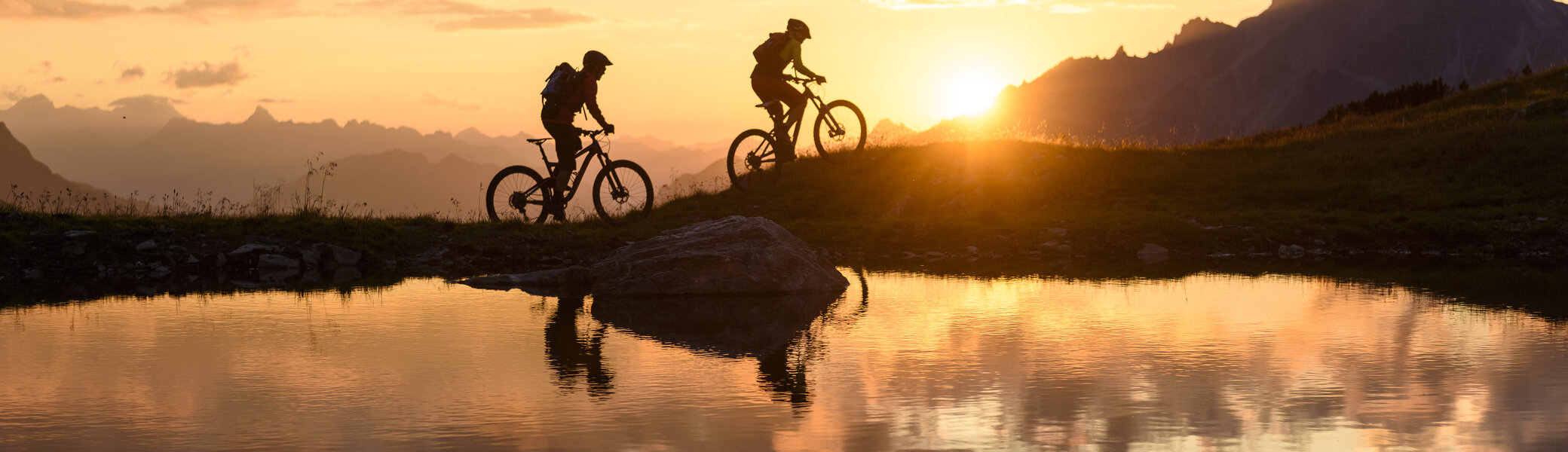 Zwei Personen fahren im Sonnenuntergang mit ihren Mountainbikes vor einer Bergkulisse. | © DAV/Wolfgang Ehn